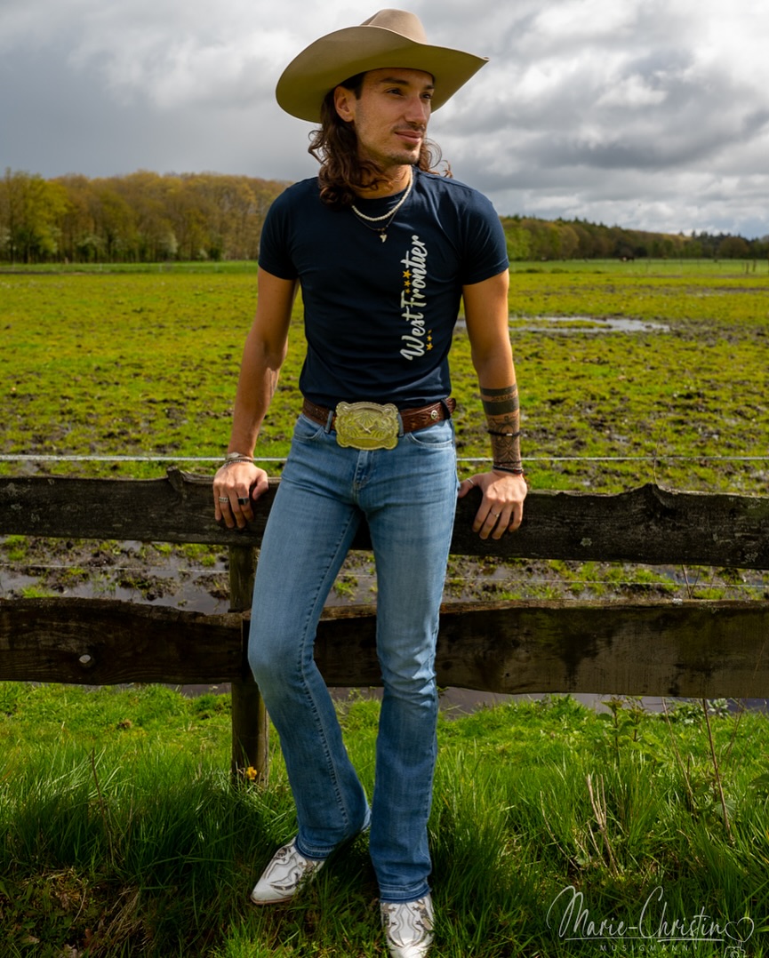 Johnny Rossato mit Cowboyhut, an einen Holzzaun auf freiem Feld gelehnt